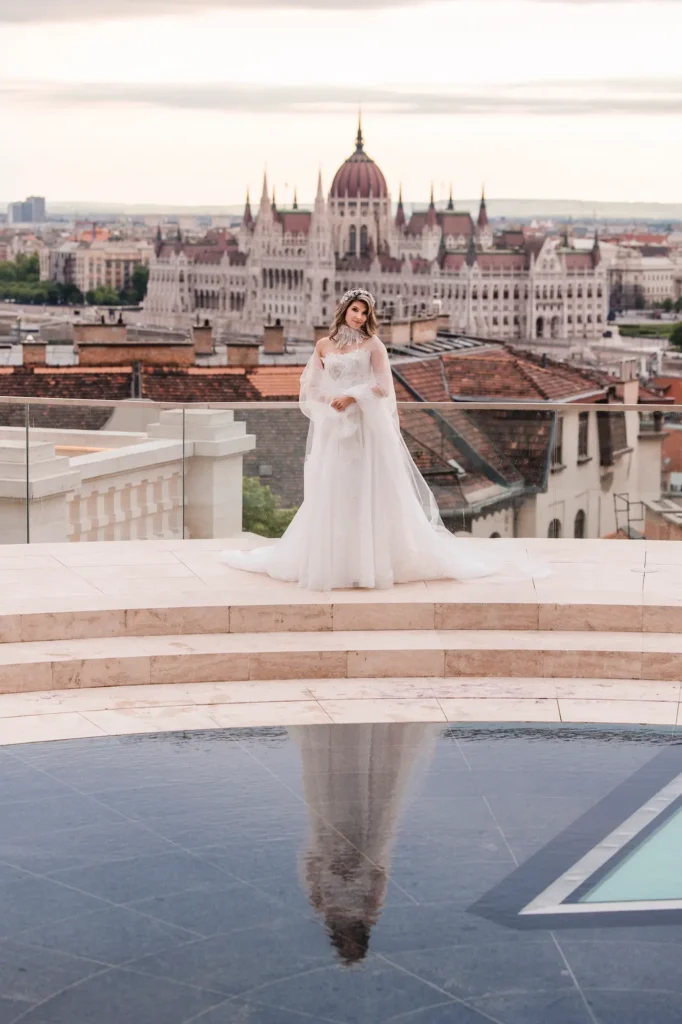 Menyasszony teljes alakos képe a Parlament panorámájával a háttérben, letisztult fehér ruhában.