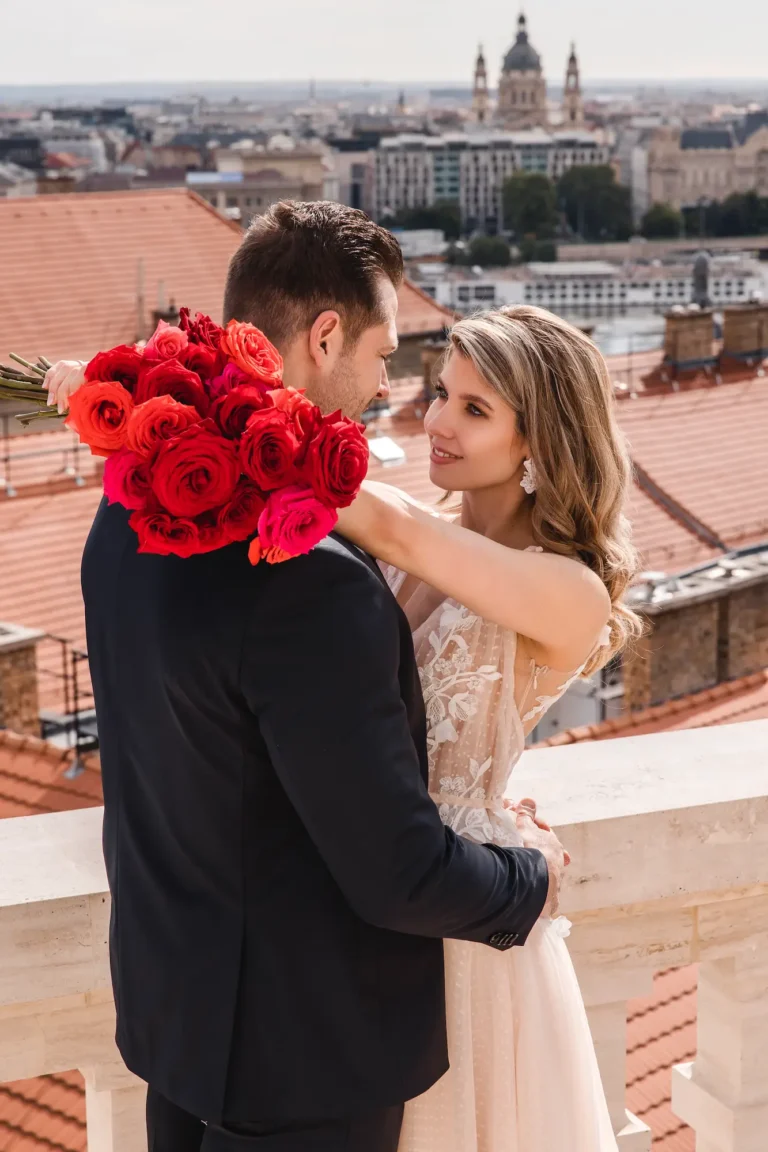 Menyasszony és vőlegény ölelkezve a budapesti tetők felett, vörös rózsacsokorral a kezükben.