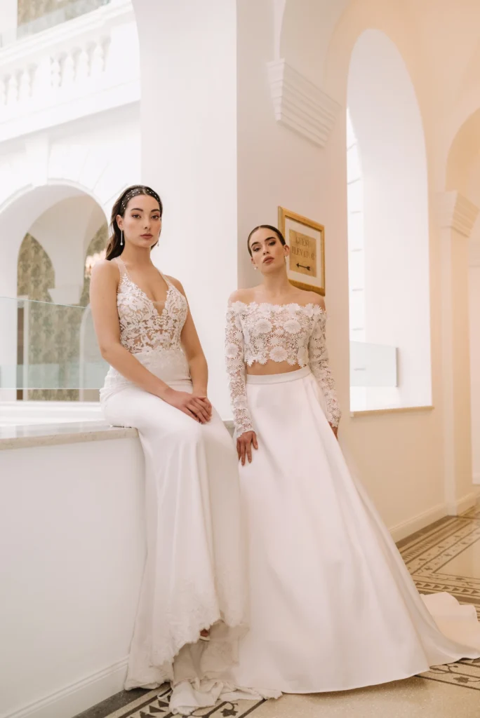 Két menyasszony modern csipkeruhában elegáns beltéri helyszínen, világos ívekkel a háttérben