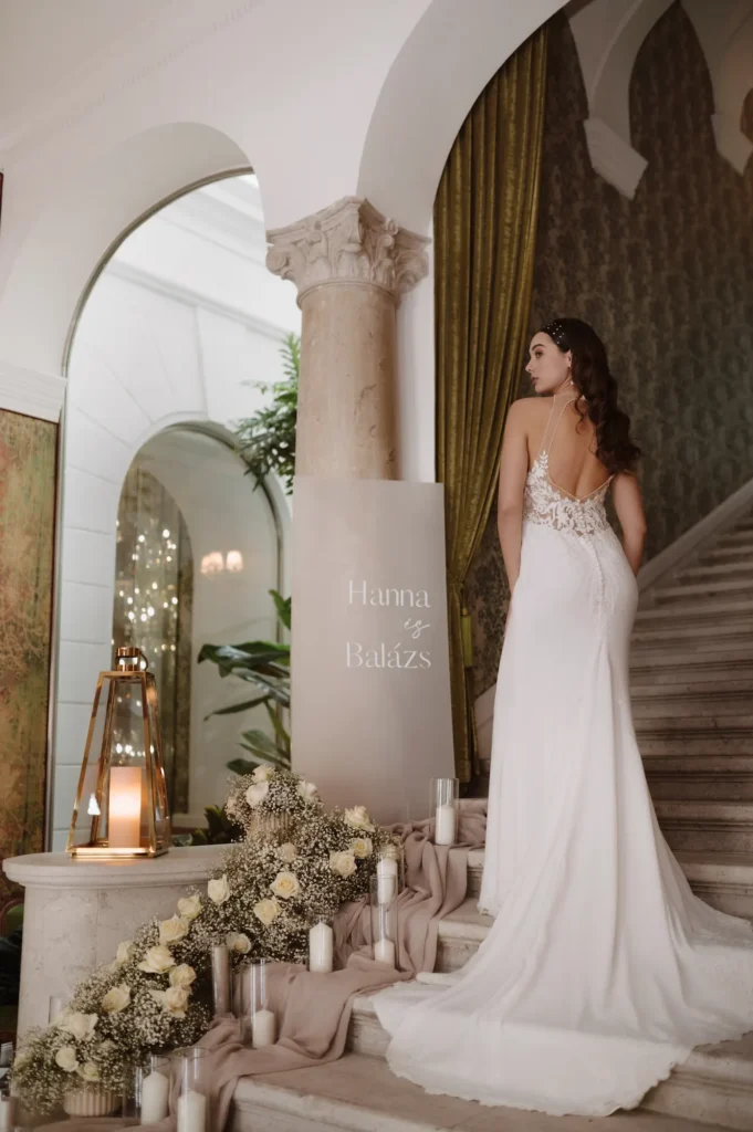 Menyasszony fehér csipkés ruhában lépcsőn áll, romantikus virágdíszítéssel és gyertyákkal körülvéve