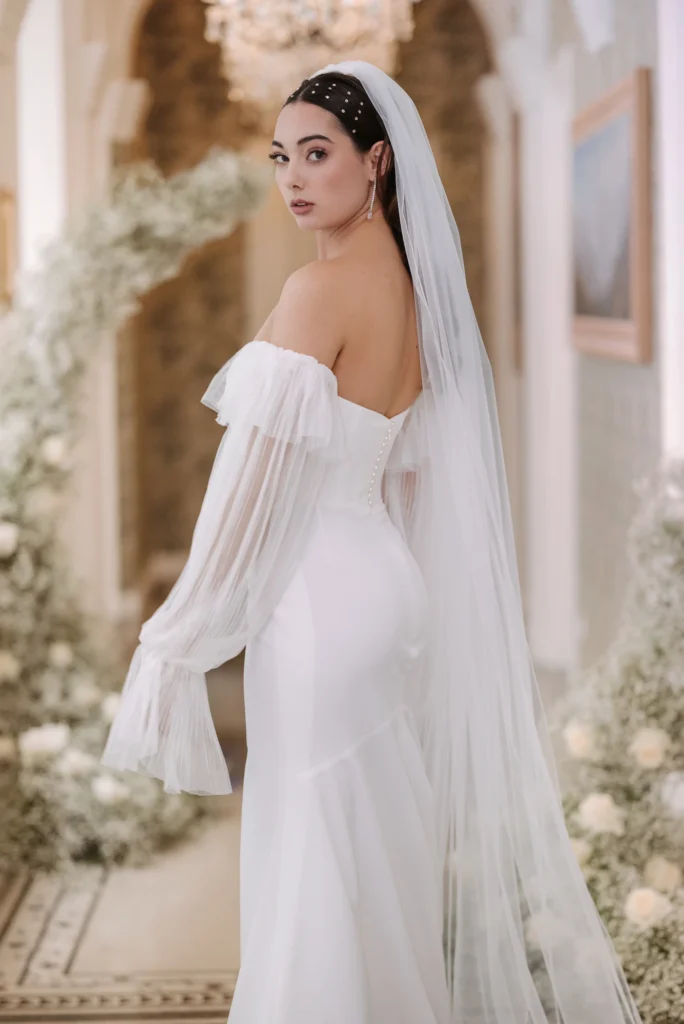 Menyasszony fátyollal és lágy tüll ujjú ruhában, klasszikus belső térben hátrafordulva