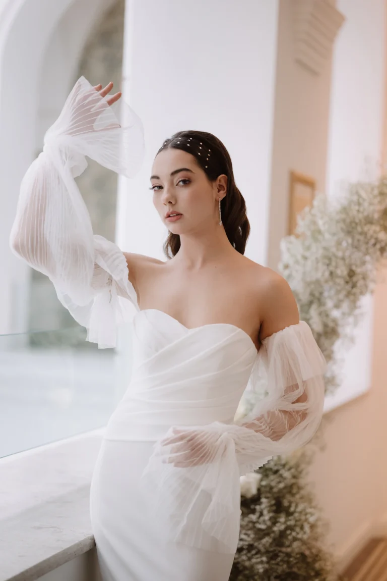 Menyasszony tüll ujjú, váll nélküli ruhában, elegáns pózban természetes fénynél